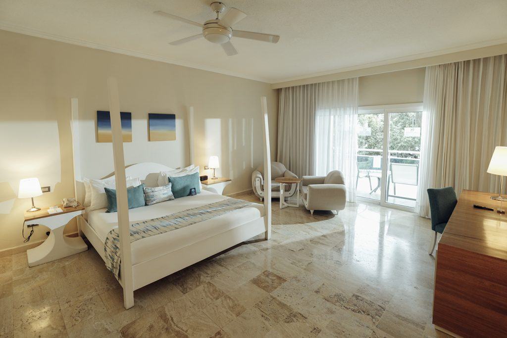 Daytime Junior Suite with a white four-poster bed, teal and blue pillows, two armchairs, terrace doors and a ceiling fan