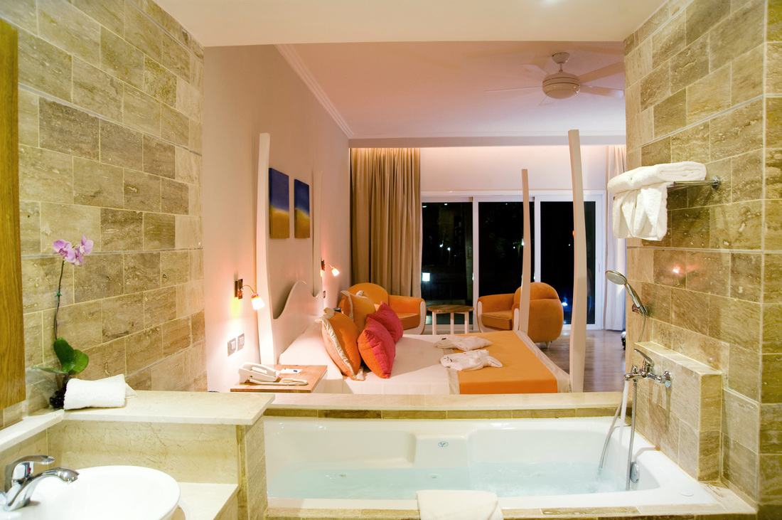 Jacuzzi tub and orchid in the stone bathroom, looking into the bedroom with orange pillows at night